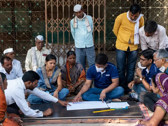 A group of people work on a project outdoors. Some of the people are standing and others are sitting. One person is writing on a a large sheet of paper on the ground. 