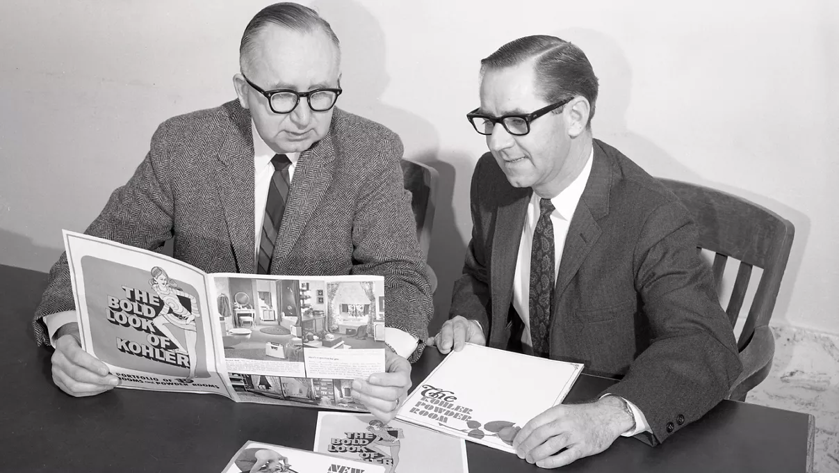 Black-and-white archives image of Bud Grube, former Communications Director for Kohler Co., sitting at a table reviewing advertising layouts with a colleague.