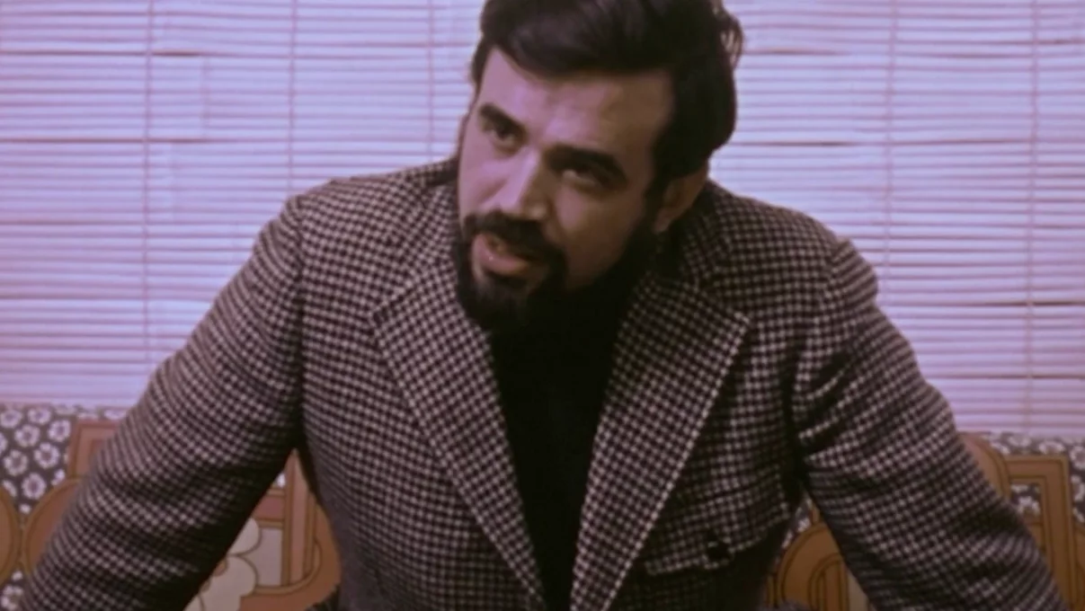 Archive image of Herb Kohler from the 1970s, sitting on a couch for an interview, looking at the camera and speaking.