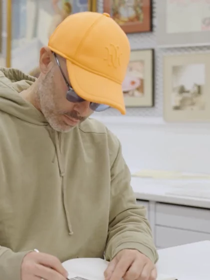 Image of global artist Daniel Arsham working on a pencil sketch of the Kohler 150th logo. He is inside the Kohler archives, looking at open reference books on a table in front of him.